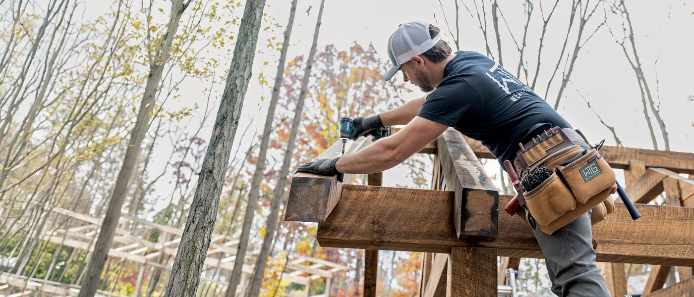 A worker wearing a leather tool belt uses a drill while building a wooden structure outdoors.